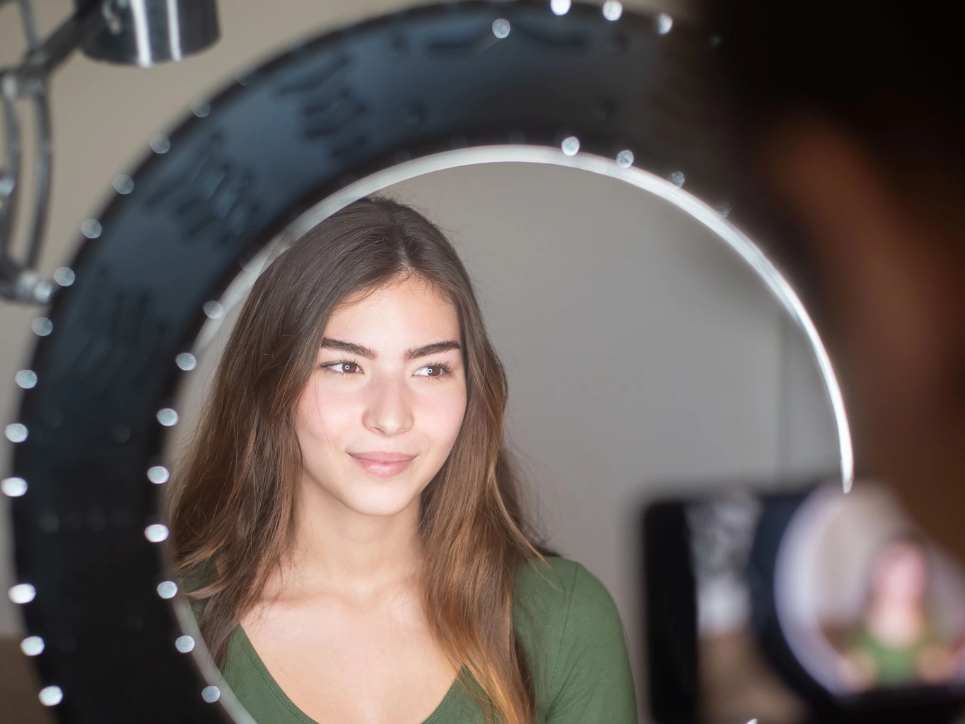 A young woman seated inside a ring light filming herself with a phone on a tripod in a home content-creator setup