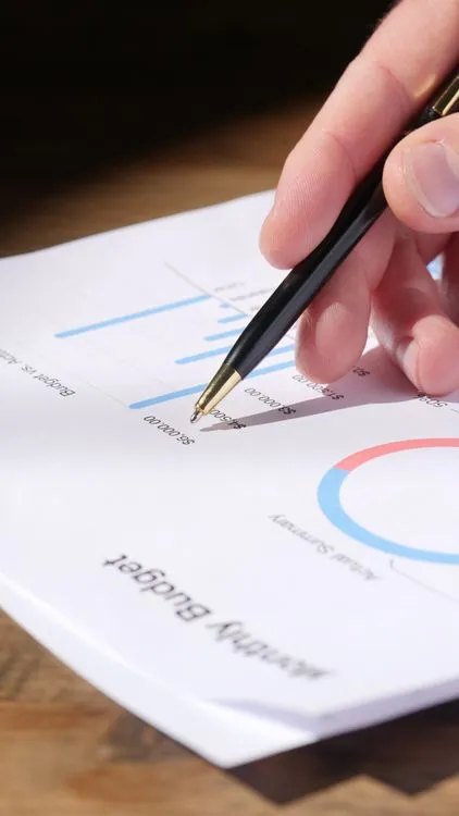 Hand with pen analyzing a monthly budget document with graphs and charts