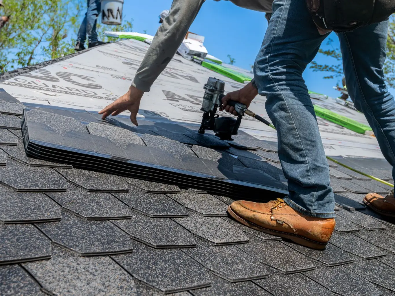 A roofer using a nail gun to install asphalt shingles on a house roof under a blue sky